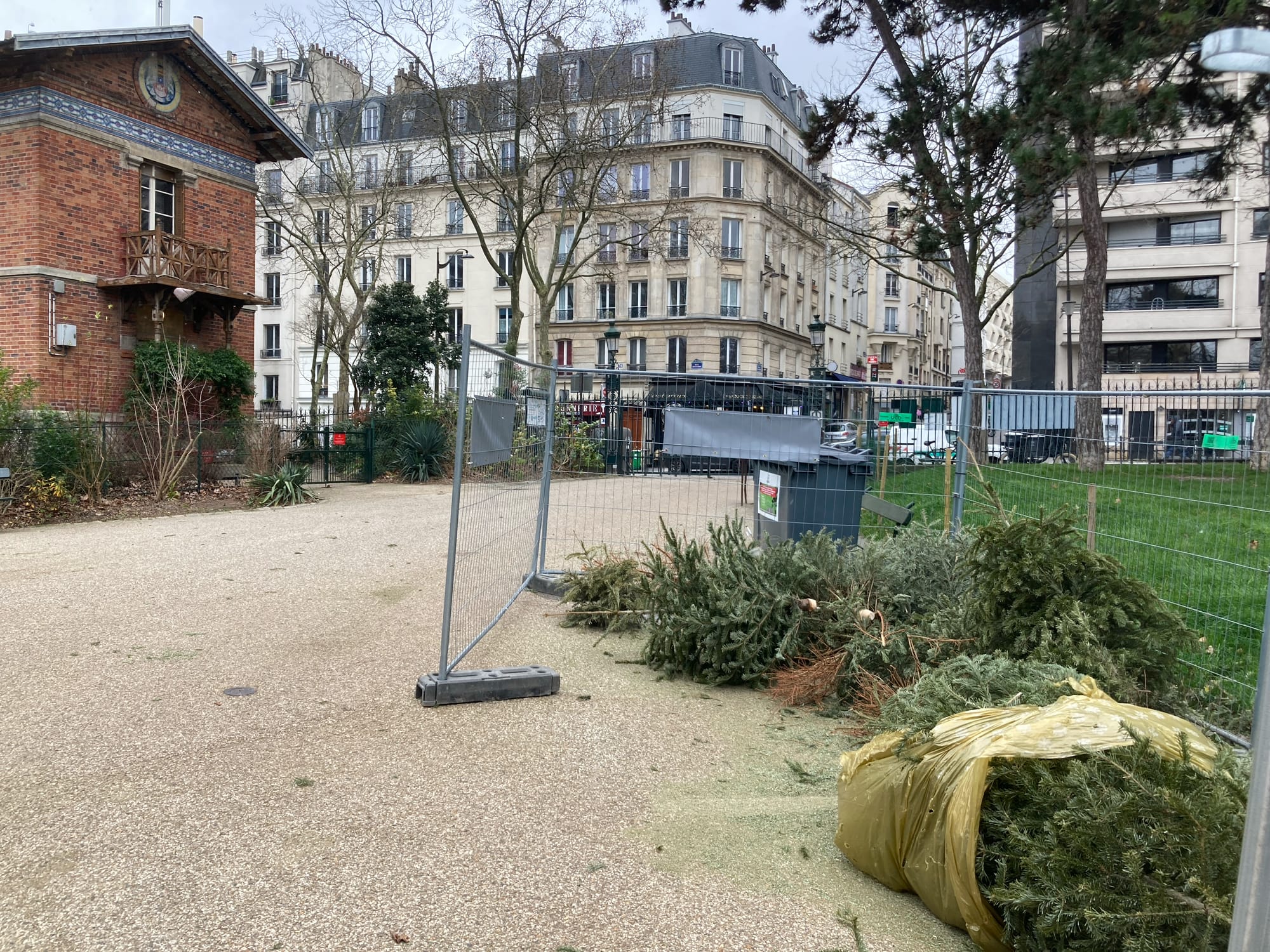 At the front of the photo is a partially fenced-in space where there are a half dozen Christmas trees are lying on their sides. To the right is a path in the Buttes Chamount park and a two storey building that would have once been the caretaker's hut in red-brick. Behind that, the photo looks onto the street and a typical Hausmann-style six storey apartment.   