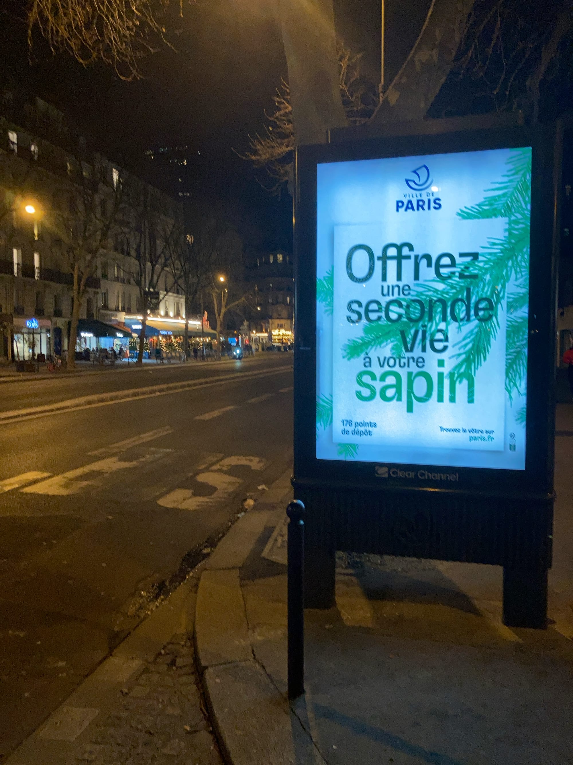 The photo is taken night, looking along a typical Parisian Street. At the roadside is a lit-up sign (one of the scrolling ones that changes to show different ads. It is sponsored by the city of Paris and says "Offrez une seconde vie à votre sapin." 